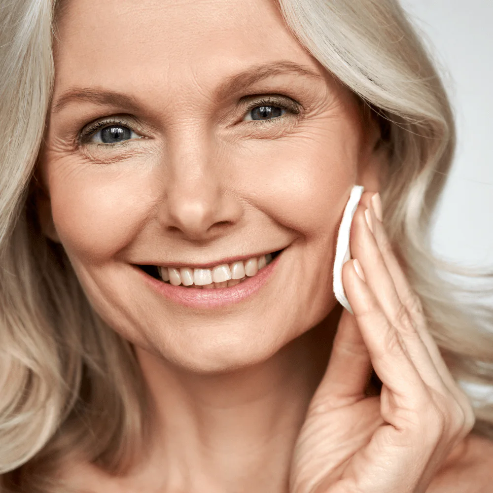 Beleznay Skincare Clinik Beleznay Évi kozmetikus - Idősebb, világos bőrű, szőke hajú, zöld szemű, barna szemöldökű mosolygós nő korongvattával törli a bőrét, természetes sminkben, szürke háttér előtt